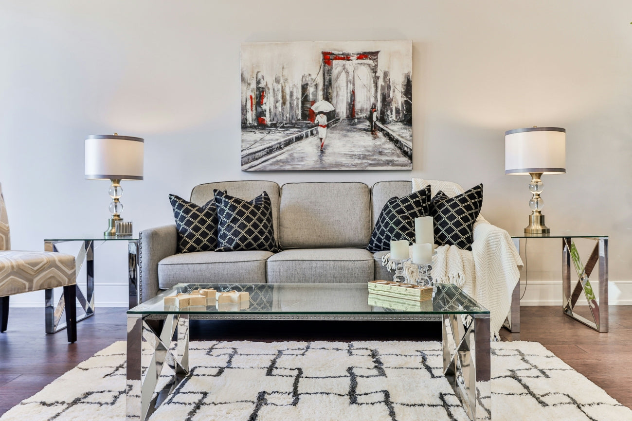 The Creation Loft – Modern living room with a gray sofa, patterned pillows, glass coffee table, elegant side tables with lamps, and contemporary wall art above the couch.